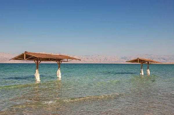 İsrail, Ein Bokek -26 Mar 2011: İki ahşap pavyon ve sazdan çatıları turkuaz Ölü Deniz 'in su bacakları İsrail sahillerinde beyaz tuz formasyonlarıyla kaplanmış Ürdün kıyılarına bakıyor. Seyahat için eşsiz doğal tuz fenomenini yakalar