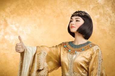 Happy smiling Egyptian woman like Cleopatra with thumbs up gesture, on golden background