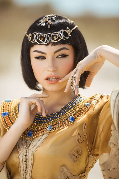 Beautiful woman with fashion make-up and hairstyle like Egyptian queen Cleopatra outdoors against desert — Stock Image Beautiful woman with fashion make-up and hairstyle like Egyptian queen Cleopatra outdoors against desert Stock Photo