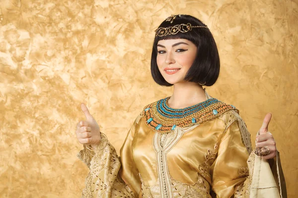 Happy smiling Egyptian woman like Cleopatra with thumbs up gesture, on golden background — Stok fotoğraf