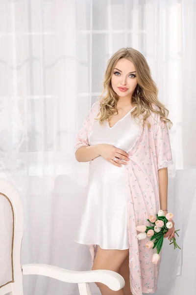 Attraktive junge blonde schwangere Frau posiert im weißen Nachthemd. glücklich Glamour sexy Mädchen zu Hause. — Stockfoto