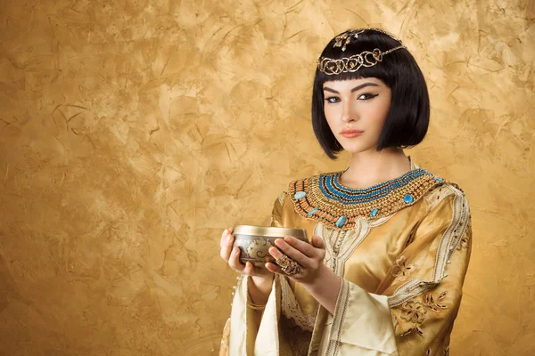 Beautiful woman like Egyptian Queen Cleopatra with cup on golden background — Stock Image Beautiful woman like Egyptian Queen Cleopatra with cup on golden background Stock Photo