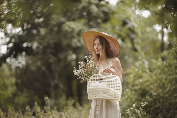 Stylish girl walking through meadow with a basket of field flowers. Young natural blonde woman with hat enjoying romantic summertime atmosphere in nature.