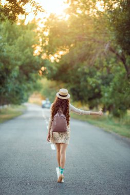 Şapkalı ve sırt çantalı bir kız yolda otostop çekiyor.