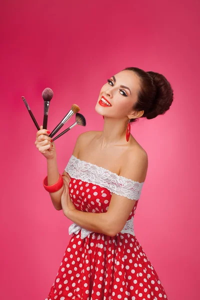 Mulher sorridente com pincéis de maquiagem. Ela está em pé contra um fundo rosa . — Fotografia de Stock