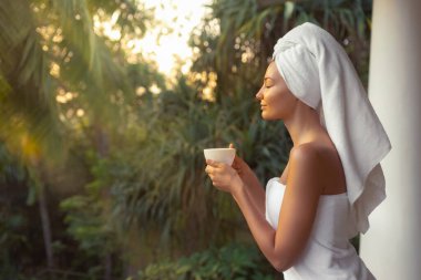 Havluya sarılıp sarılarak kahveyi yudumlayan güzel bir kadın. Parlak güneş ışığı altında tropikal balkonda gözleri kapalı, spa, rahatlama, dinlenme, dinlenme, tatil ve kendine dikkat..