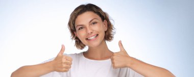 Beyaz tişörtlü neşeli genç bir kadın mavi arka planda çifte onay veriyor. Reklamlar, olumlu mesajlar, marka görselleri veya motivasyon promosyonları için mükemmel.