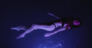 Mor tonlu loş bir havuzda yüzen bir kadının stilize edilmiş dikey görüntüleri. Güzellik tanıtımları, moda görselleri veya yaratıcı ticari kullanım için ideal sanatsal konsept.