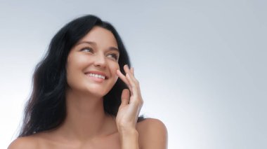 Yüzüne hafifçe dokunurken neşeyle gülümseyen açık tenli, koyu renk saçlı beyaz bir kadın. Cilt bakımı, güzellik ürünleri, sağlık ve yüksek kaliteli kozmetik reklamları için harika..
