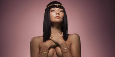 Düz siyah saçları ve altın mücevherleri olan şık bir kadın yumuşak pembe bir arka plana karşı güvenle poz verir. Onun çıplak omuzları ve sakin ifadesi onun zarafetini ve lüksünü vurguluyor. Tam oldu.