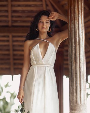 Beyaz, sırt dekolteli yaz elbisesi giyen beyaz, beyaz bir kadın, elinde çiçeklerle, tahta bir çardakta güvenle duruyor. Güzellik, moda, kaplıca ve romantik yaşam tarzı kavramları için harika..