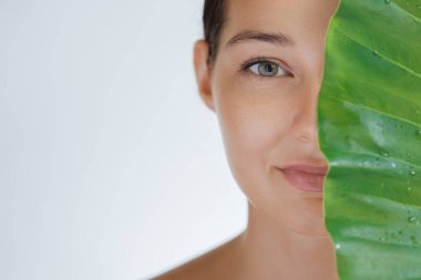Parlayan derisi kısmen tropikal yaprağın arkasında su damlalarıyla gizlenmiş bir kadının banner tarzı görüntüsü. Cilt bakımı, eko güzellik, sağlık, su ya da kozmetik tedavi için ideal.