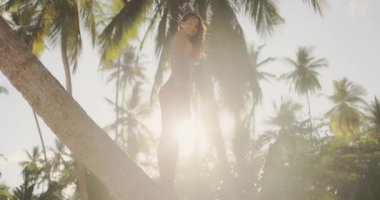 Güneş ışınları tropikal yeşilliklerden süzülen eğimli bir palmiye ağacının üzerinde duran kırmızı bikinili güzel bir model. Yaz, seyahat ve egzotik yaşam tarzı terfileri için mükemmel.. 