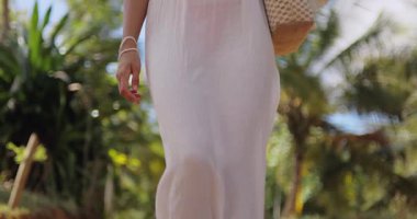 Hasır şapka ve beyaz yaz kıyafeti giymiş mutlu bir kadın tropikal bir ortamda duruyor, parlak güneşin altında gülümsüyor. Rahatlama, seyahat ve yaşam tarzı temaları için ideal. 