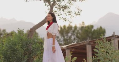 Beyaz elbiseli, kırmızı eşarplı şık bir kadın ağaçların ve dağların yanında yürüyor. Yaşam tarzı, seyahat, moda tanıtımı, reklam ve doğa için mükemmel..