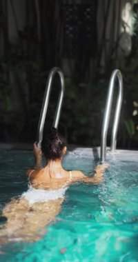 Beyaz mayo giymiş, turkuaz bir tatil köyünde yüzen ve tırmanan bir kadının sakin sahnesi. Seyahat, tatil, spa ve tropikal yaşam tarzı kavramları için ideal.