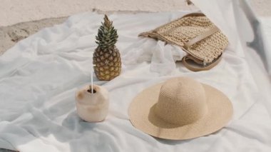 Bir hasır şapka, ananas, hindistan cevizi içeceği ve el yapımı bir çadırın altında beyaz bir kumaş üzerine örülmüş dokuma poşeti içeren sakin bir plaj sahnesi. Yaz, tatil ya da yaşam tarzı kavramları için mükemmel. 