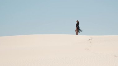 Çölde kum tepeciğinde duran bir kadının ağır çekim videosu. Siyah pelerini rüzgarda zarifçe çırpınıyor. Güzelliği, özgürlüğü ve doğanın gücünü yakalar..