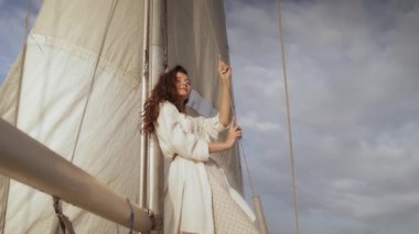 Yatta duran beyaz elbiseli zarif bir kadın elinde yelkenle dramatik bir gökyüzünün altındaki mesafeye bakıyor. Lüks seyahat, macera ve yaşam tarzı terfileri için mükemmel.. 