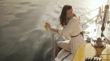 Beyaz elbiseli güzel bir kadın bir yatın kenarında oturuyor, bir yaprak tutuyor ve altın gün batımının altında sakin okyanusun tadını çıkarıyor. Lüks yaşam tarzı ve seyahat promosyonları için ideal.. 
