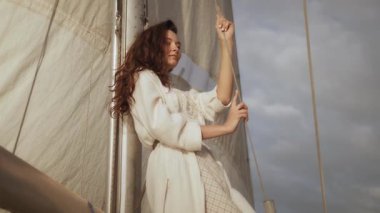 Beyaz elbiseli şık bir kadın yatta yelkenli tutuyor, rüzgarın ve dramatik bulutların tadını çıkarıyor. Lüks yaşam tarzı kampanyaları, seyahat promosyonları ve deniz maceraları için ideal.. 