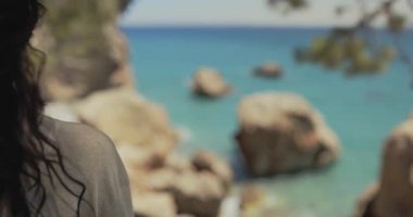 Kayalık kıyısı olan turkuaz okyanusa bakan bir kadının arkasından güzel bir yavaş çekim. Kamera geniş manzaralı. Seyahat, yaşam tarzı ve doğa temalı görüntüler için ideal.