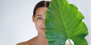 Cilt bakımı ve sağlık pankartıyla bronzlaşmış bir kadın büyük yeşil bir yaprağın arkasında su damlacıklarıyla gülümsüyor. Çevre dostu güzellik, spa hizmetleri, organik kozmetik ürünleri ve cilt suyu reklamları için harika..