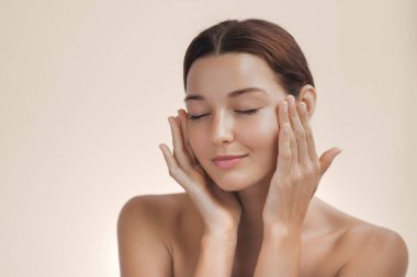 Beyaz tenli ve doğal makyajlı genç bir kadının kaliteli portresi. Yüzüne nazikçe kapalı gözlerle dokunuyor. Cilt bakımı, sağlık, rahatlama ve kaplıca tedavisi kavramı