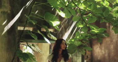 Beyaz elbiseli çarpıcı bir kadın, canlı tropik yeşilliklerle çevrili antik harabelerde zarifçe poz veriyor, içinden güneş ışığı akıyor. Seyahat veya moda görselleri için ideal. 