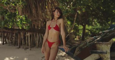 Kırmızı bikinili genç bir kadın uzak bir Sri Lanka plajında kırsal bir ahşap teknenin yanında güven içinde poz veriyor. Yaz aylarında moda kampanyaları, plaj kıyafetleri reklamları ya da egzotik seyahat görselleri için mükemmel.. 