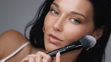 Genç bir kadının, kusursuz cildini ve ışıl ışıl güzelliğini vurgulamak için fırçayla makyaj yaptığı detaylı bir yakın çekim. Güzellik, cilt bakımı ve kozmetik reklamlar için ideal..