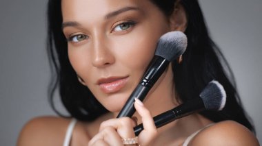 İki makyaj fırçası tutan çarpıcı bir kadın çok yönlü kozmetik aletlerin önemini vurguluyor. Dekoratif kozmetik, güzellik dersleri ve ürün seçimleri için ideal..