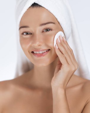 Kafasında havlu olan güzel bir kadın yüzünü temizlemek için pamuk pedi kullanırken gülümsüyor. Cilt bakımı, cilt bakımı ve güzellik anlayışı sağlıklı, parlayan ciltler için.. 