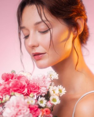 Parlak tenli, doğal makyajlı, elinde karanfil ve papatya buketi olan genç bir kadının zarif bir stüdyo resmi. Güzellik, cilt bakımı, sağlık ve çiçek temalı markalar için ideal..