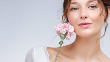 Stüdyoda, yüzünün yanında pembe bir karanfil tutan zarif, beyaz bir kadın. Kusursuz bir cilt, yumuşak bir makyaj ve sakin bir görünüş. Güzellik, cilt bakımı ve kozmetik markalar için ideal..