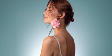 Parlak tenli ve omuzunda pembe karanfil olan zarif bir genç kadının hayali stüdyo portresi. Cilt bakımı, çiçek, sağlık, moda ve romantik temalı promosyonlar için ideal..