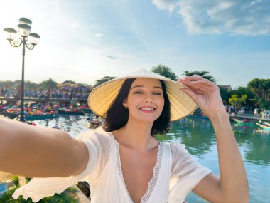 Geleneksel şapkasız genç kadın gülümseyip Vietnam 'da Hoi An' da renkli nehir teknelerinin yanında selfie çekiyor. Parlak seyahat tarzı imajı bloglar, turizm ve sosyal medya reklamları için mükemmel.