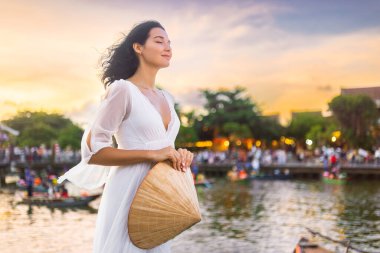 Beyaz elbiseli zarif kadın Vietnamlı şapkasını elinde tutuyor ve güneş batarken Hoi An nehrinin kıyısında saçında rüya, yaşam tarzı, moda ya da seyahat içeriği için ideal..