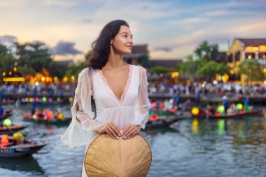 Hoi An, Vietnam 'da fenerle aydınlatılmış nehrin kenarında duran güzel kadın, rüzgar saçlarını hafifçe kaldırırken elinde konik bir şapka tutuyor. Barışçıl, romantik bir seyahat imajı yaşam tarzı ve kültürel kampanyalar için mükemmel..