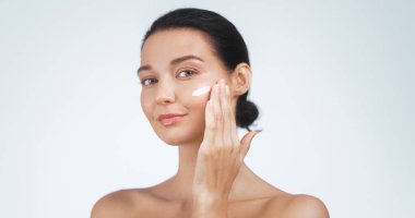 Yanağına nazikçe yüz kremi süren beyaz bir kadının stüdyo görüntüsü. Cilt gösterimi rutin ve parlak bir cilt. Güzellik reklamları, ürün ambalajları ve cilt tanıtımları için mükemmel..