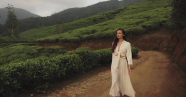 Sri Lanka 'daki yemyeşil çay tarlaları arasında kırsal bir yolda yürüyen beyaz kıyafetli zarif bir kadının yavaş çekim videosu. Yaşam tarzı, seyahat, moda ve kültür turizmi için ideal.