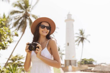 Sri Lanka 'da palmiye ağaçları olan Galle Deniz Feneri' nin önünde şapka ve güneş gözlüğü takan neşeli genç kadın, tropik tatil, gezi fotoğrafçılığı ve macera kavramı.