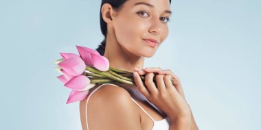 Omzunda pembe nilüfer çiçekleri tutan Asyalı bir kadının profili güzellik ve huzuru sembolize ediyor. Cilt bakımını, kaplıcayı ve doğal zarafeti teşvik etmek için ideal.