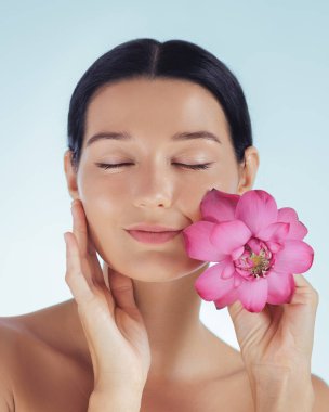 Güzel, genç bir kadın. Gözleri kapalı ve yanağında pembe nilüfer çiçekleri var. Yüzünde sakin bir ifade ve parlak bir ten var. Kozmetik reklamlar, cilt bakımı ve doğal sağlık görselleri için ideal..