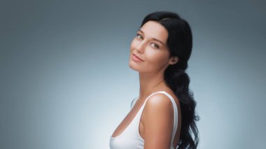 Beyaz atlet bluzuyla hafifçe gülümseyen açık tenli, koyu renk saçlı beyaz bir kadının stüdyo portresi. Cilt bakımı, yaşam tarzı ve sağlıkla ilgili promosyon içeriği için ideal.