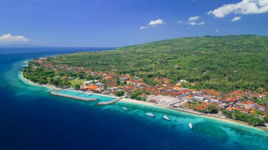 Nusa Penida 'daki Sampalan Limanı' nın canlı havası dalgalı bir dalgakıran, feribot iskelesi ve mercan mavisi bir kıyı şeridi ve çatıları olan sürat tekneleri gösteriyor. Bali seyahati ve turizm reklamları için yüksek kaliteli görüntü.