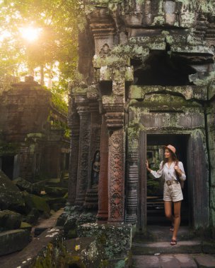 Ta Prohm Angkor Wat Kamboçya Tapınağı 'nın altın saatinde, şapkalı ve sırt çantalı beyaz bir kadın yosunlu Khmer kapısından içeri giriyor. Turizm, kültür ve editoryal kullanım için yüksek kaliteli seyahat imajı.