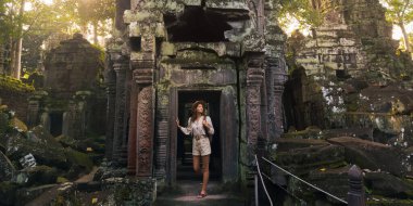 Ta Prohm Angkor Wat Kamboçya Tapınağı gün doğumunda parlayan, şapka ve sırt çantalı beyaz bir kadın oyulmuş kapı aralarını ve yosunlu taşları keşfediyor. Editoryal kullanım için yüksek kaliteli seyahat resmi.