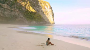 Hafif elbiseli bir kadının rüya gibi yavaş çekim sahnesi. Kelingking Sahili 'nde dalgaların yanında oturuyor. Nusa Penida, Bali..