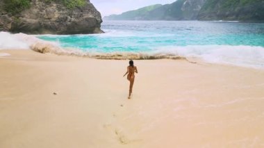 Ağır çekimde Kelingking Beach, Nusa Penida, Bali 'deki turkuaz kıyı şeridine doğru neşeyle koşan bir kadın görülüyor. Seyahat reklamları, yaşam tarzı temaları ve yüksek kaliteli tropikal içerik için ideal..